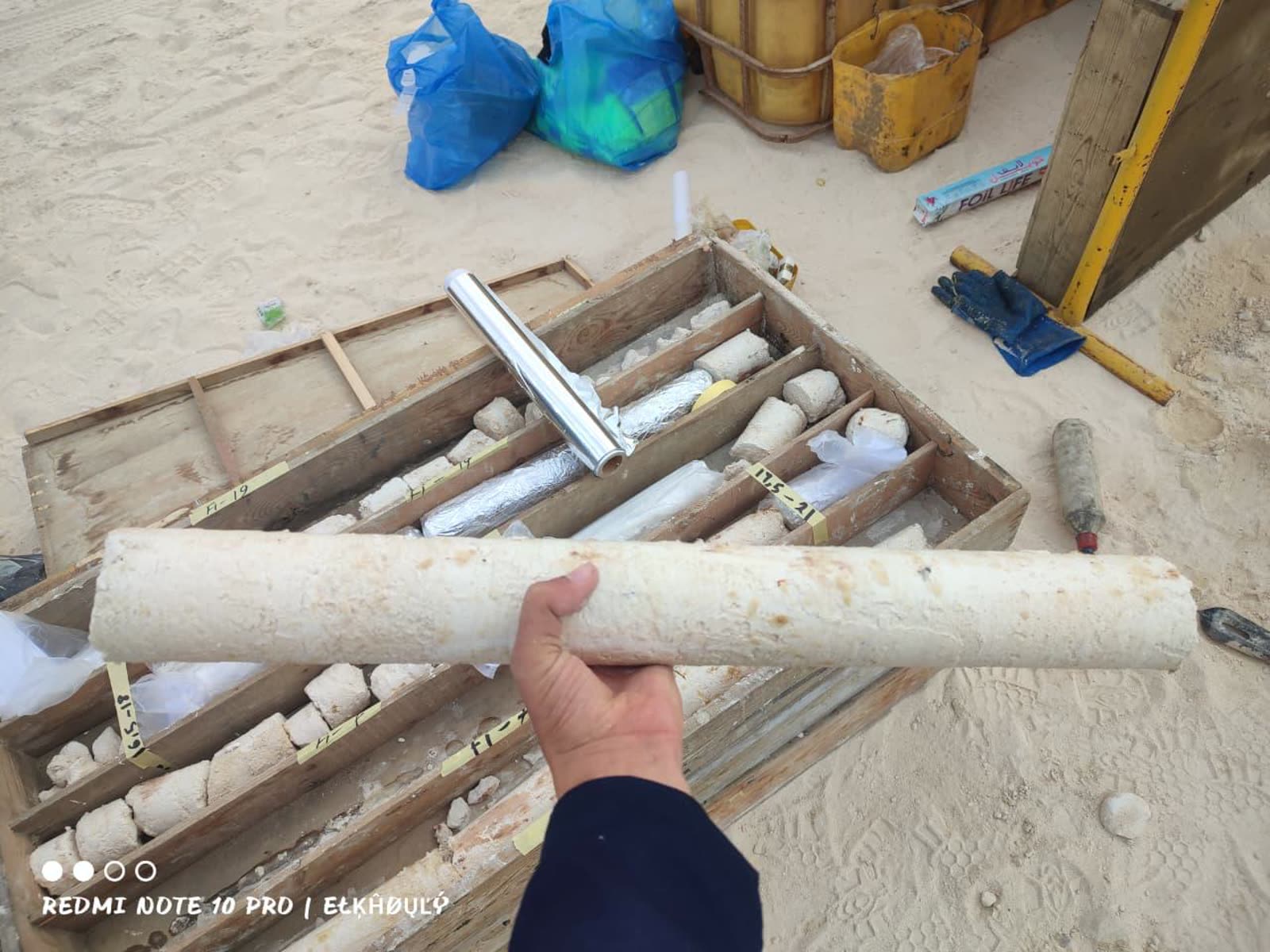 Horema technician holding a recovered limestone rock core beside a labelled core box of samples on a coastal site