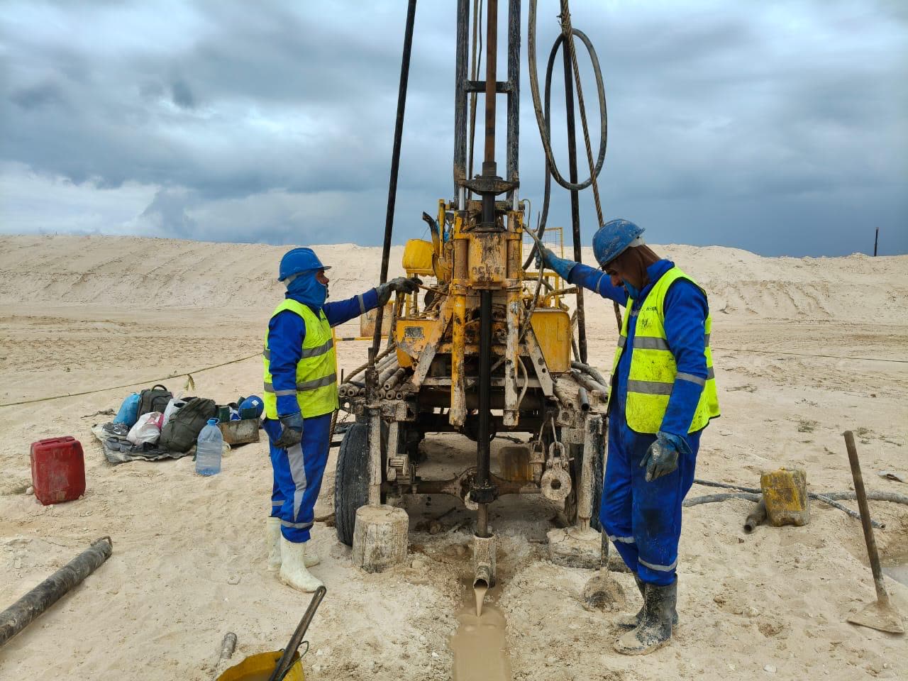 Two Horema drillers working a rotary rig under a heavy storm sky on a sand site